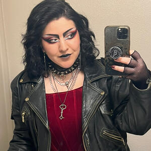 a selfie of me. i have wavy black hair, my makeup is done in a trad goth style, and i’m wearing a black leather jacket and red bodice-style top.